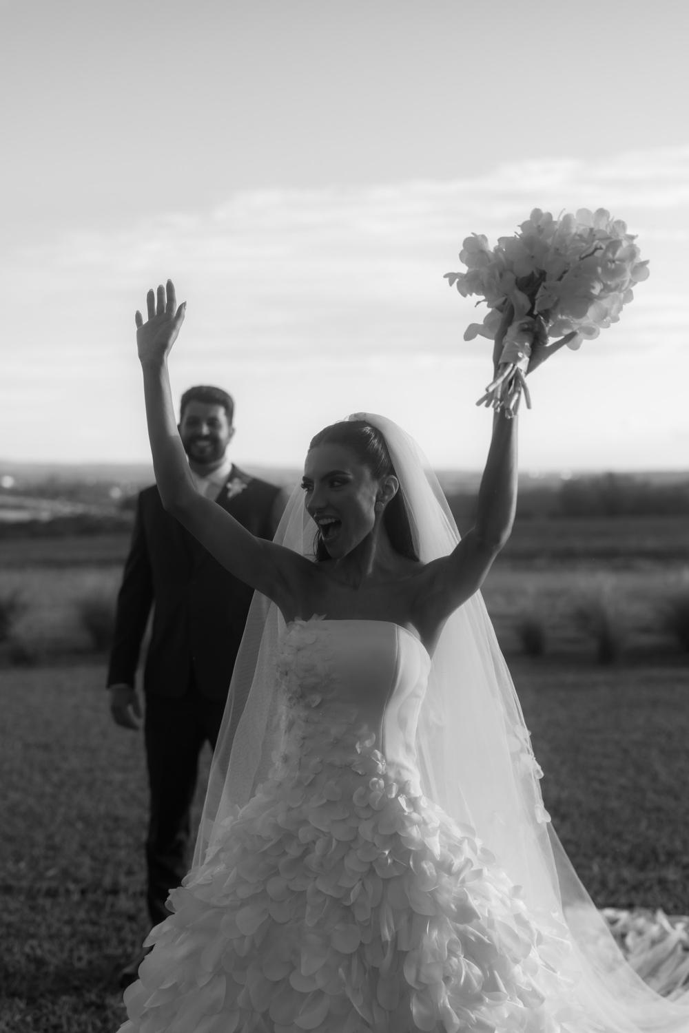 Bride celebrating with her arms raised and the bouquet, groom smiling behind her